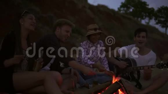 一群年轻快乐的人晚上坐在海边的火炉旁烤香肠弹吉他视频的预览图