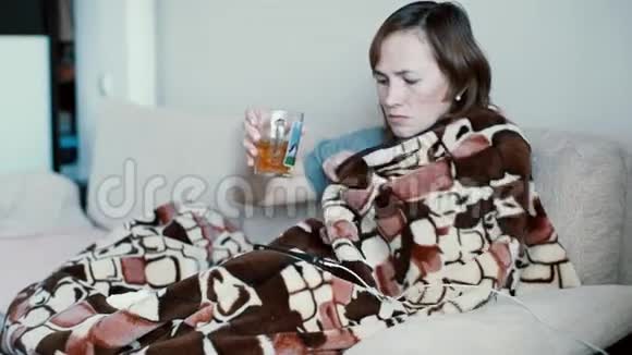 生病的女人在家里坐在沙发上盖着毯子喝着温暖的饮料视频的预览图