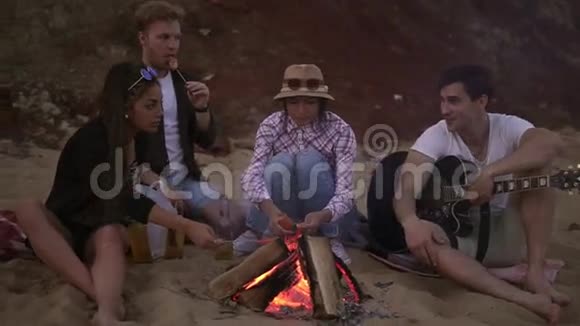 一群年轻快乐的人晚上坐在海边的火炉旁烤香肠弹吉他视频的预览图