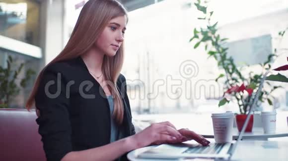 穿着黑色夹克的金发女孩坐在咖啡馆A里她喝了一杯咖啡用笔记本工作视频的预览图