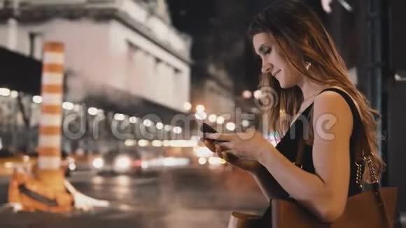 年轻漂亮的女人晚上站在市中心在美国纽约使用智能手机视频的预览图