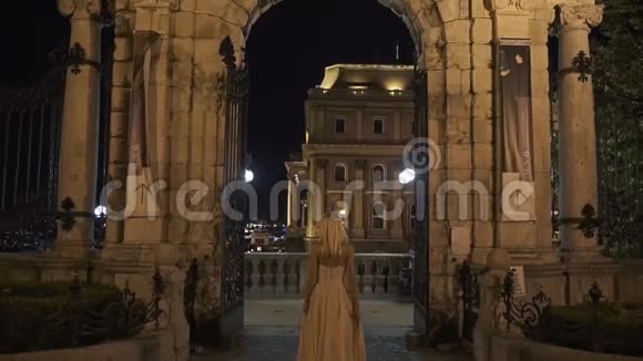 晚上在老大理石城堡的阳台上摆姿势的穿着优雅粉红色长裙的漂亮女士的后景视频的预览图