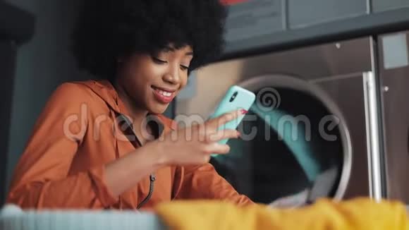 在洗衣店用智能手机微笑的年轻女人自助公共洗衣店视频的预览图