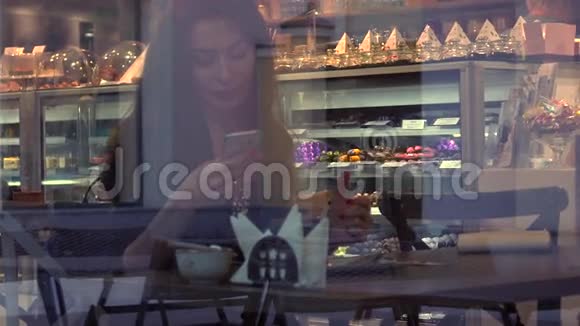 漂亮的黑发女孩在咖啡馆里用她的智能手机从外面透过窗户观看4K视频视频的预览图