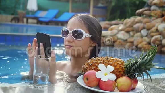 年轻的美女在游泳池旁边的水杯和带热带水果的盘子里使用手机视频的预览图