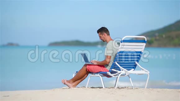 带笔记本电脑的年轻人在加勒比海热带海滩坐在日光浴床上用电脑在海滩上工作的人视频的预览图