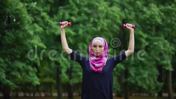 身穿罩袍的年轻穆斯林妇女在夏季公园里用哑铃做运动视频的预览图