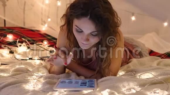 年轻女子躺在床上床上放着一块放在花环背景上的平板电脑在网上购物视频的预览图