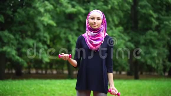 身穿罩袍的年轻穆斯林妇女在夏季公园里用哑铃做运动视频的预览图
