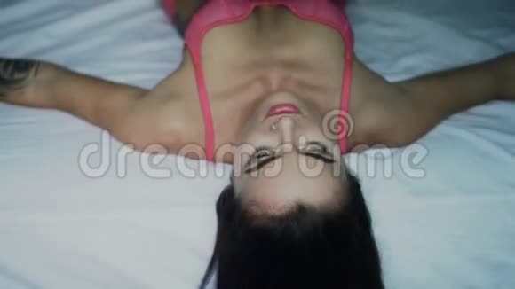 年轻美丽的黑发女人穿着粉红色内衣躺在床上微笑着欣赏着早晨视频的预览图