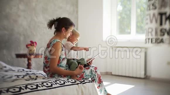 年轻的妈妈带着婴儿看着平板电脑带着阳光在房间里微笑视频的预览图