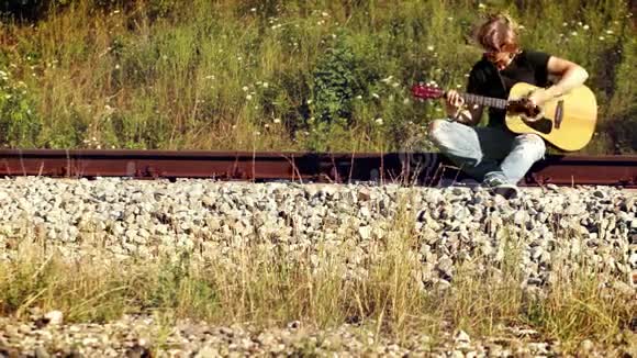 在阳光明媚的日子里金发的吉他手坐在铁路上演奏视频的预览图