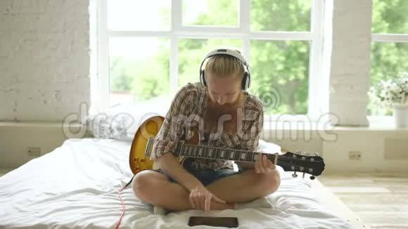 现代卧室里戴着耳机坐在床上学习用平板电脑弹吉他的漂亮胡子男视频的预览图