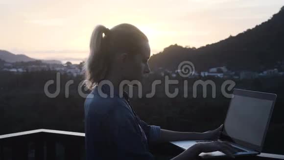 一个漂亮的金发女人站在美丽的日出的屋顶上用笔记本电脑工作视频的预览图