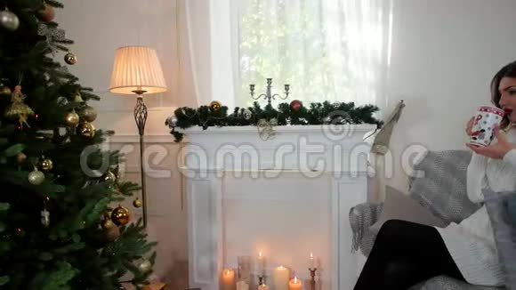 一位年轻女子喝茶的肖像在圣诞树附近的客厅里快乐的女孩喝茶微笑的样子视频的预览图