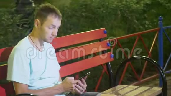 有魅力的悲伤男人坐在一张带智能手机的长凳上在温暖的夏夜在公园里4kStealam射击视频的预览图