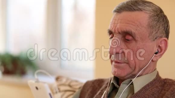 戴着白色智能手机的老人坐在沙发上听音乐视频的预览图