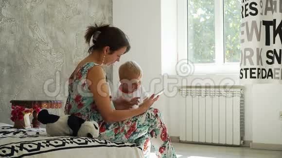 年轻的妈妈带着婴儿看着平板电脑带着阳光在房间里微笑视频的预览图