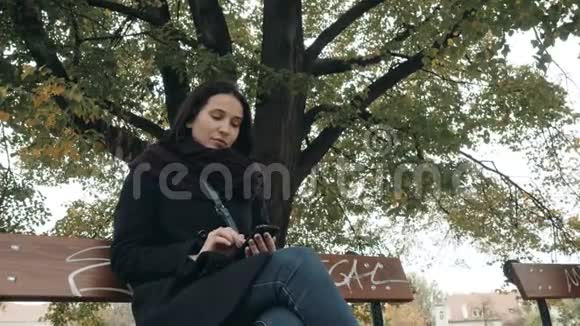 年轻的微笑妇女使用智能手机坐在公园的长凳上美丽的欧洲女孩打电话发短信视频的预览图