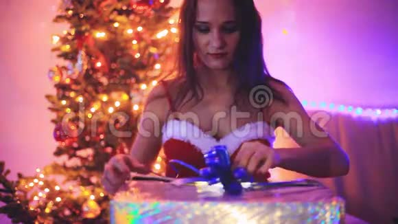 年轻快乐的女人打开神奇的圣诞礼物手持闪亮的灯光女孩坐在圣诞树旁边装饰视频的预览图
