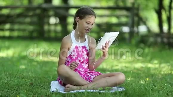 美丽的女孩穿着粉红色的衣服带着平板电脑坐在花园里的绿草上视频的预览图