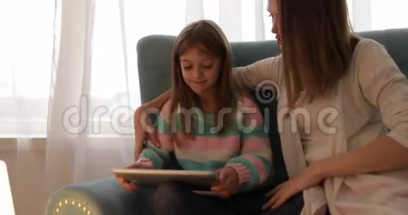 母亲和女儿坐在沙发上用平板电脑快乐地微笑着年轻的家庭视频的预览图