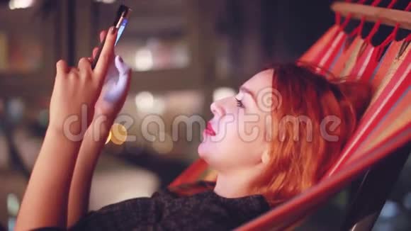 美丽的女人红头发在城市里使用智能手机晚上躺在吊床上1920x1080视频的预览图