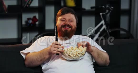 一个长着胡子的胖子笑着在房间里看电视在桌上吃爆米花和啤酒视频的预览图
