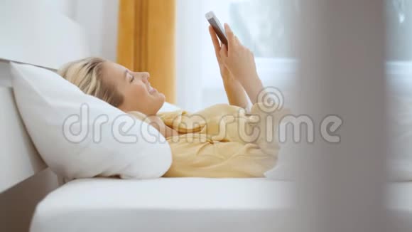 年轻的金发女人躺在床上打电话视频的预览图