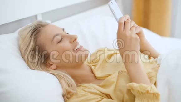 年轻的金发女人躺在床上打电话视频的预览图