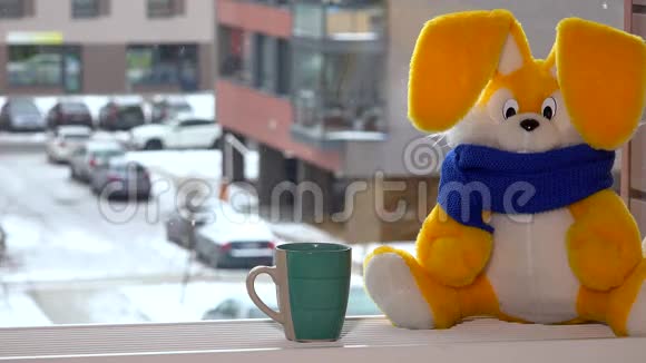冬天黄色玩具兔子拿着茶杯坐在窗边雪花飘落视频的预览图