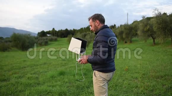 一个长胡子的男人穿着羽绒服看着他的平板电脑一边微笑着在田野里驾驶无人机视频的预览图