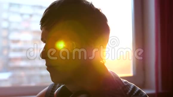 一个年轻人站在窗户附近戴上耳机听音乐背景模糊与日落伙计视频的预览图