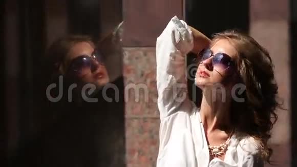 迷人的年轻微笑的女人戴着太阳镜穿着白色衬衫在大理石附近的阳光下站着卷着的小鸽子视频的预览图