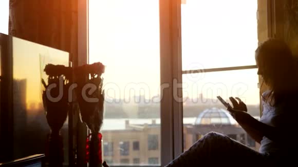 坐在窗户上的漂亮年轻女人在日落和城市的背景下使用智能手机3840x2160视频的预览图