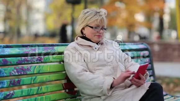 女人坐在秋色城市公园的长椅上使用红色智能手机视频的预览图