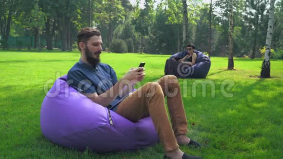一个穿着蓝色衬衫的年轻人坐在公园的椅子上在电话里得到了一条信息视频的预览图