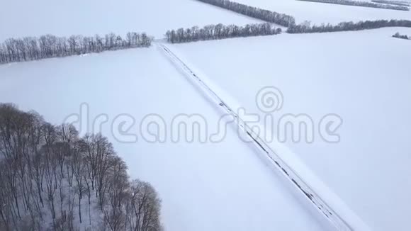 在白雪覆盖的田野之间的冬季森林道路上行驶的汽车的鸟瞰图视频的预览图