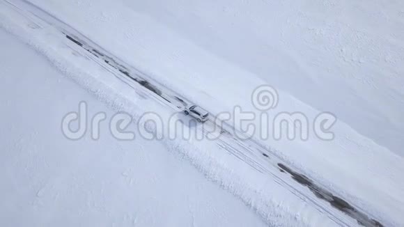在白雪覆盖的田野之间的冬季森林道路上行驶的汽车的鸟瞰图视频的预览图
