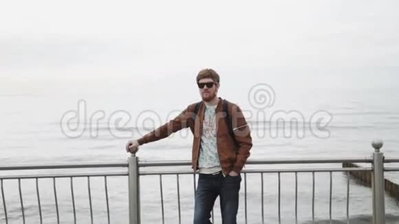 一个留着红胡子的时髦年轻人穿着棕色皮夹克戴着太阳镜背着背包微笑着视频的预览图
