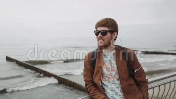 一个留着红胡子的时髦年轻人穿着棕色皮夹克戴着带背包的太阳镜微笑着视频的预览图