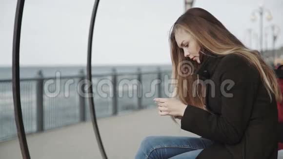 在俯瞰大海的木板路上的长凳上戴着白色智能手机的女人视频的预览图
