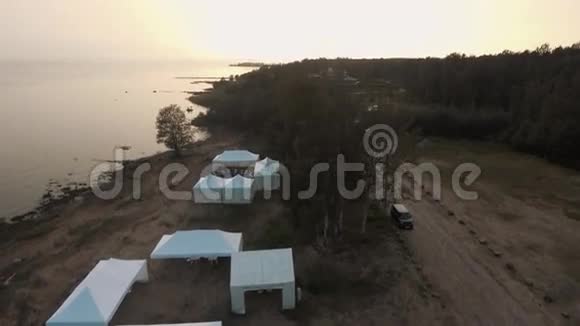 在日落时在森林附近的沙滩上可以看到白色帐篷和建筑物视频的预览图