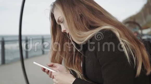 在俯瞰大海的木板路上的长凳上戴着白色智能手机的女人视频的预览图