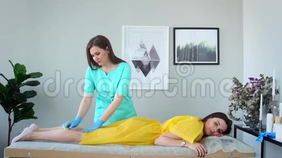 两个女孩在一家美容院主人穿着浴衣戴着手套做着用糖去除腿上毛发的程序视频的预览图