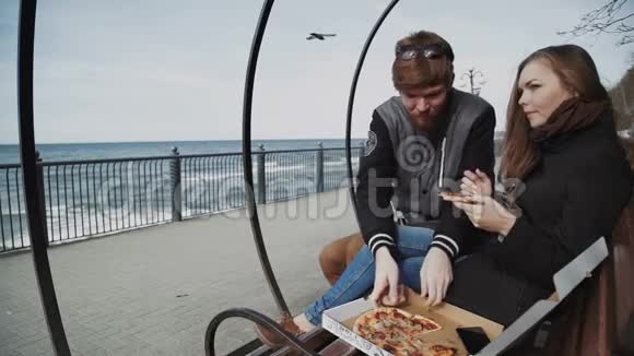 一对夫妇在海滨吃披萨天气寒冷在一起年轻人穿着夹克女人穿着外套坐在长凳上视频的预览图