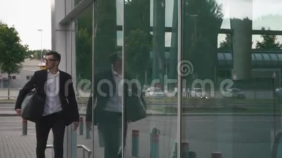 年轻自信的商人戴着眼镜带着手提箱在现代机场航站楼的玻璃墙上视频的预览图