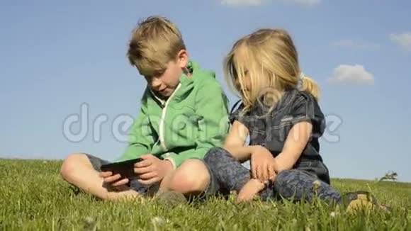 快乐的金发孩子使用智能手机看电影或玩游戏坐在草地上视频的预览图