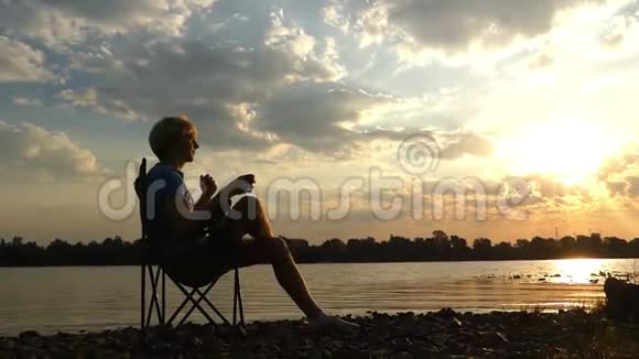 日落时分年轻人坐在河岸边喝咖啡视频的预览图