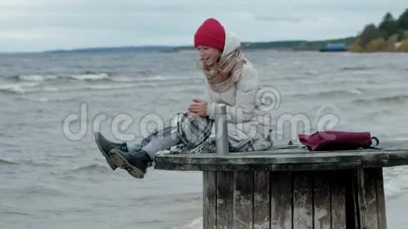 年轻女子穿着温暖的衣服坐在海边坐在木盘管上喝着热水瓶里的热茶寒冷的天气暴风雨视频的预览图